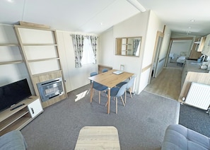 A modern, well-lit room with a flat-screen TV, a wooden dining table with chairs, a built-in fireplace, and a kitchen area in the background.