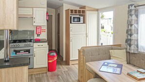 A compact kitchenette with a stove, oven, and microwave, a small trash bin, and a seating area with a table and cushions. - 2 bedroom accommodation in Gwespyr, Nr Prestatyn, Denbighshire (Gwespyr, Nr Prestatyn, Denbighshire)