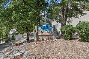 A sign with a beach scene and the text "The Beach" in front of a building with trees and a gravel path. - Ledges Landing – Waterfront Condo, Beach, & Pools! (Osage Beach)