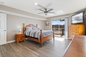 A bedroom with a wooden bed, a nightstand, a dresser, a ceiling fan, and a balcony with a view. - Ledges Landing – Waterfront Condo, Beach, & Pools! (Osage Beach)