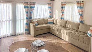 Una sala de estar con un sofá seccional color beige, una mesa de comedor dispuesta con platos, vasos y cubiertos, y cortinas a rayas.