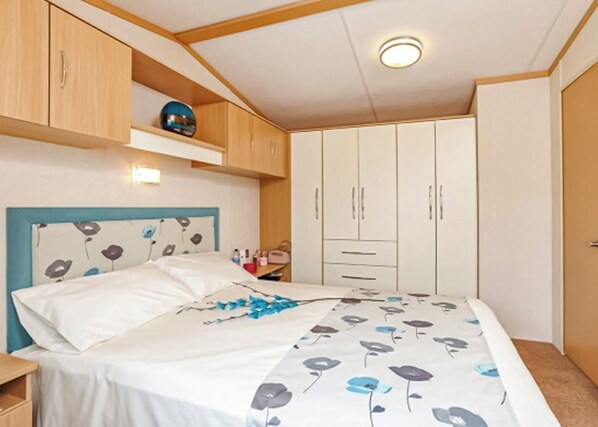 A bedroom with a bed, a nightstand, a wardrobe, and a shelf with a decorative item. - 12ft-wide caravan with one double and one twin. (Silloth)
