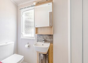 A bathroom with a toilet, a sink, a window, and a wooden cabinet. - Spacious and luxurious 12ft-wide caravan with one double and two twin bedrooms. (Axminster)