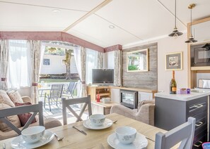 A cozy living area with a TV, dining table, and chairs, a kitchen with a microwave, and a balcony with curtains. - Spacious and luxurious 12ft-wide caravan with one double and two twin bedrooms. (Axminster)