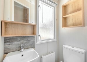 Un baño con lavamanos blanco, estantes de madera, una ventana con vidrio satinado y un radiador.