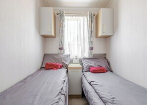 Pieni huone, jossa on kaksi erillistä sänkyä, ikkuna, jossa on verhot, ja pieni pöytä, jossa on lamppu.