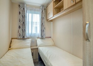 A compact room with two beds, a window with curtains, a small cabinet, and a power outlet.