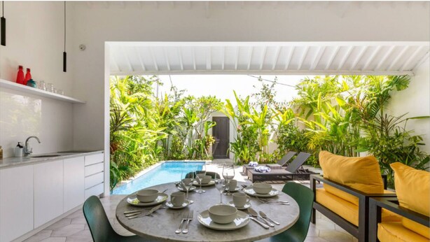 A dining area with a round table set for four, surrounded by green chairs. A pool is visible in the background, and there are plants and outdoor furniture. - Villa Harris (Canggu)