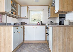 Uma cozinha compacta com armários brancos, piso em padrão herringbone e uma janela com persianas.