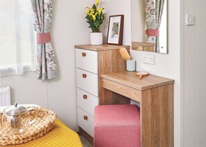 A bedroom with a wooden desk, a mirror, a bed with a yellow blanket, a potted plant, and a framed photo.