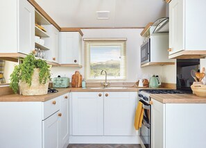 Une cuisine compacte avec des meubles blancs, un plan de travail en bois, un four encastré et une fenêtre équipée de stores.