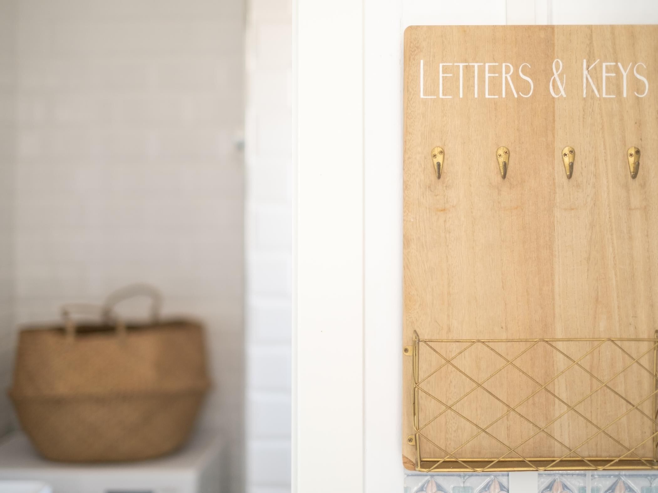Un panel de madera con la inscripción 'Letters & Keys', cuatro ganchos, una bolsa tejida y una celosía metálica en una pared.