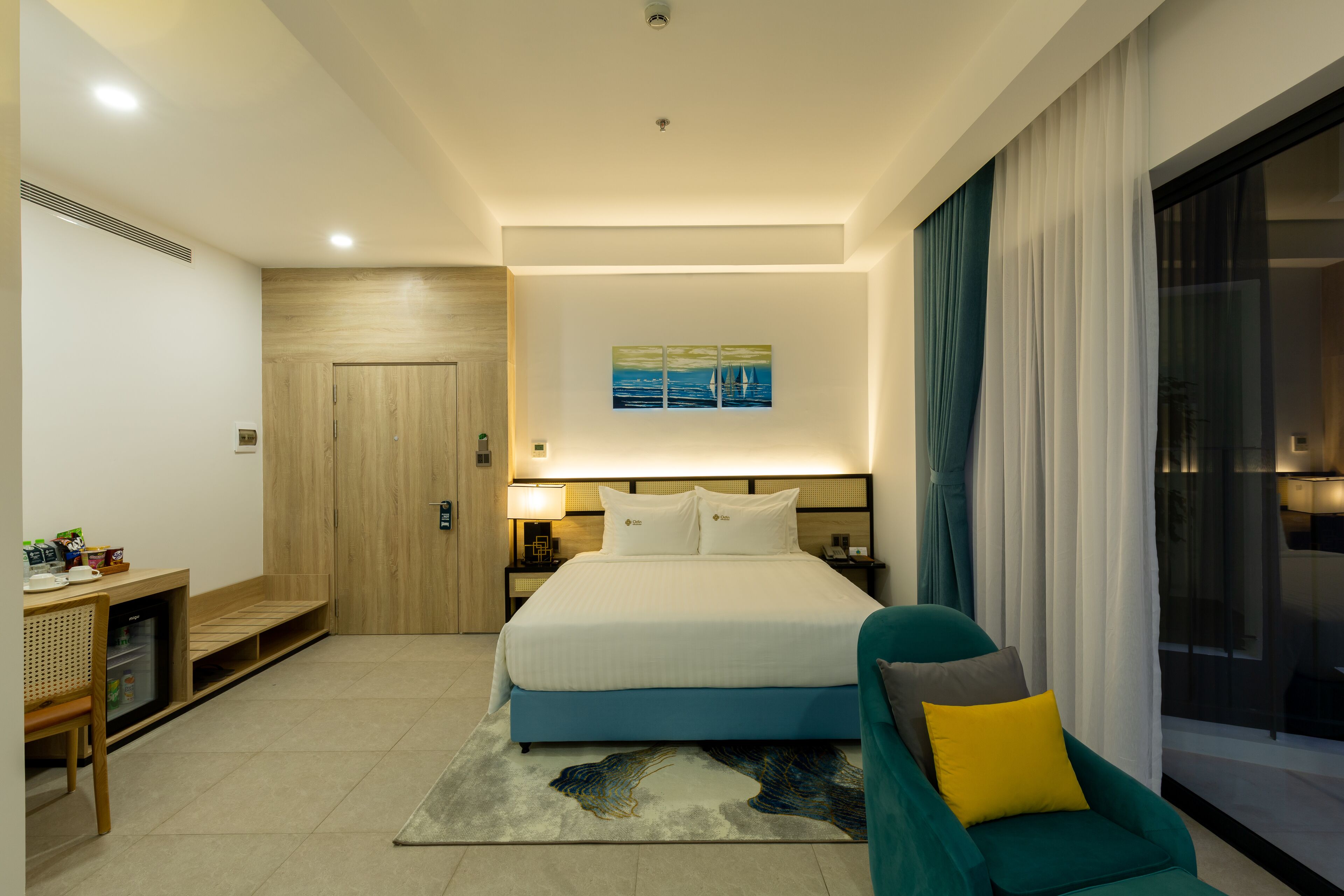A modern hotel room with a large bed, a desk with a refrigerator, a chair, and a large window with curtains.