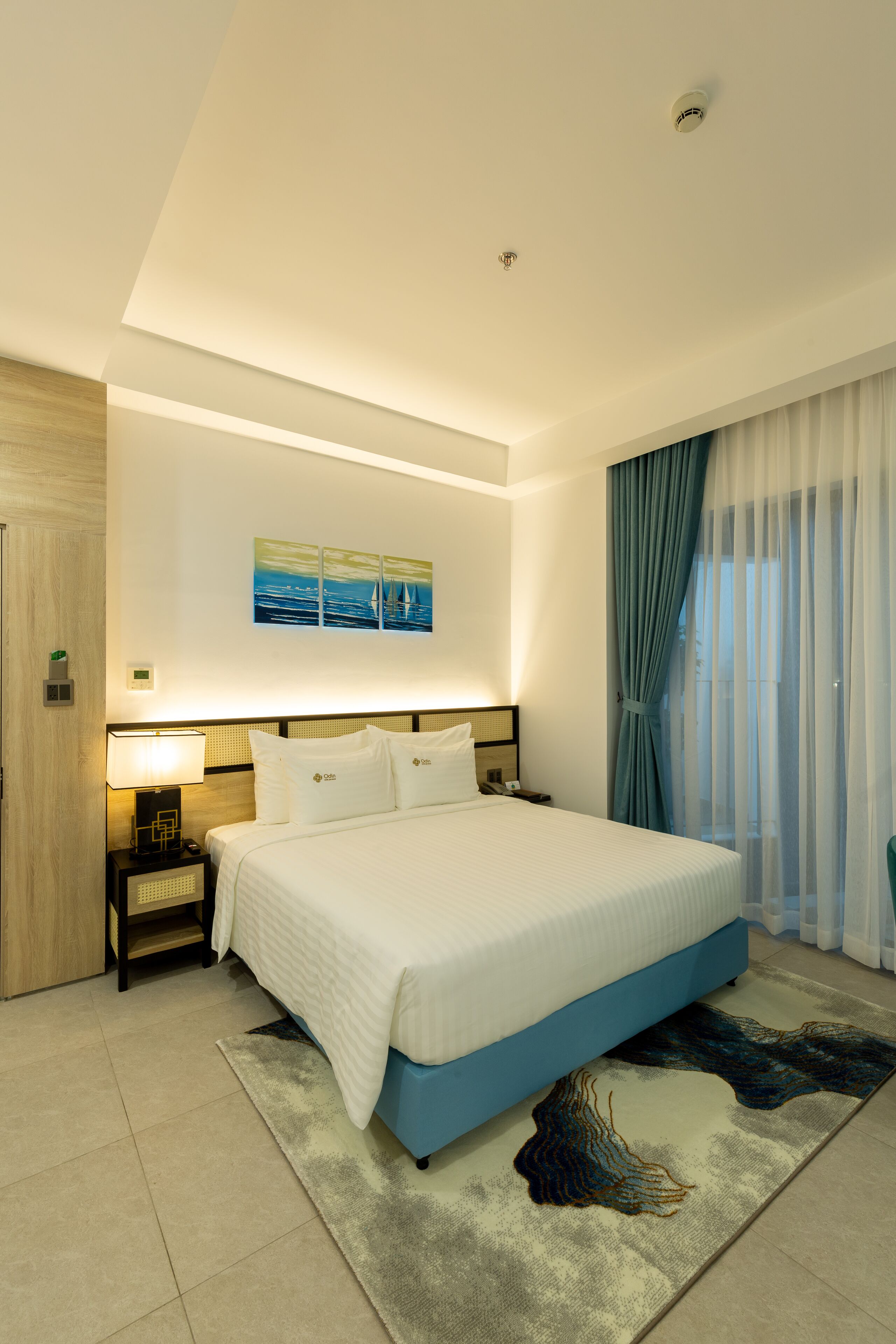 Habitación de hotel moderna con una cama grande, una mesita de noche, una lámpara de pared y vistas al exterior a través de cortinas translúcidas.