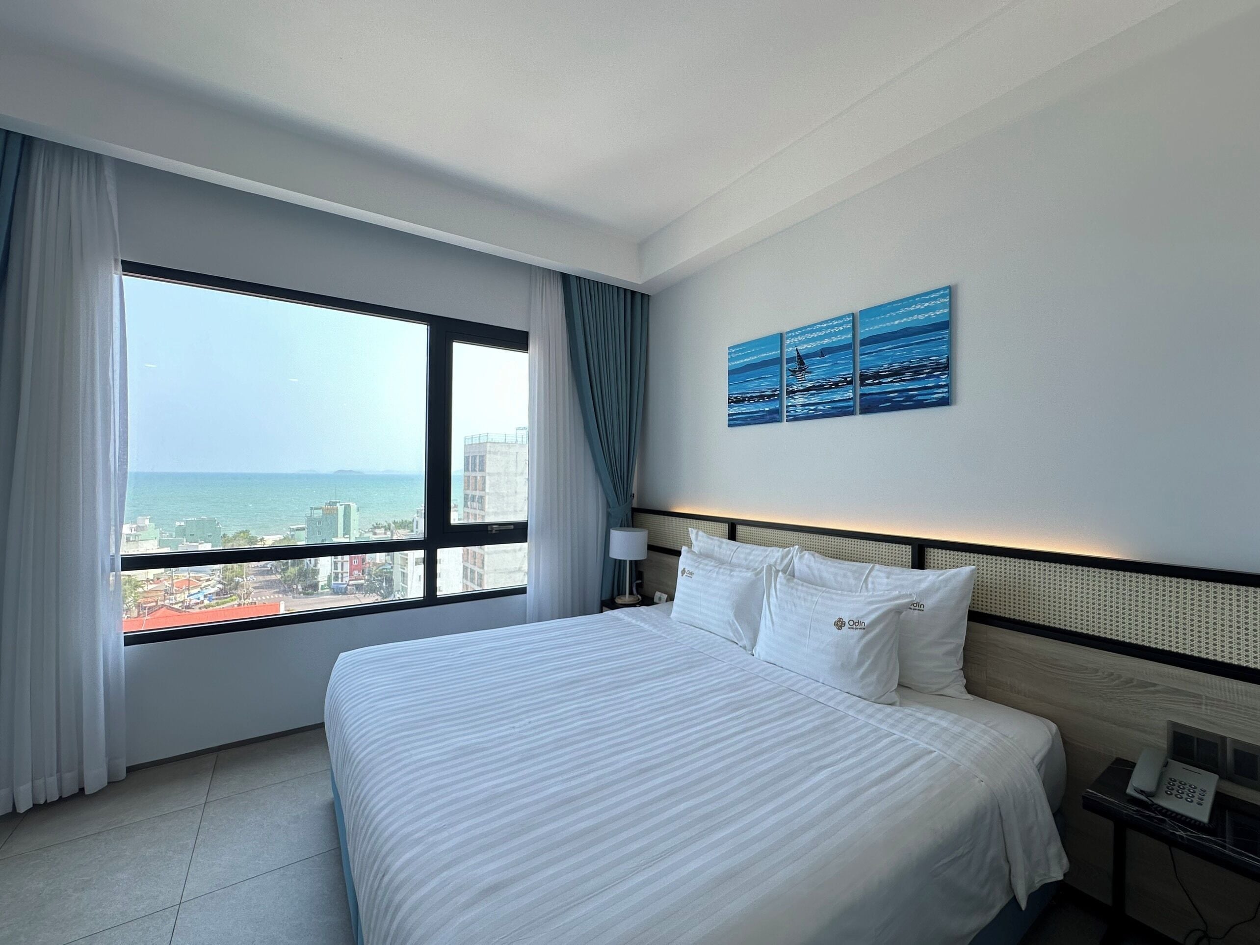 A hotel room with a large window, a bed with white bedding, a nightstand with a phone, and a wall with a blue artwork.