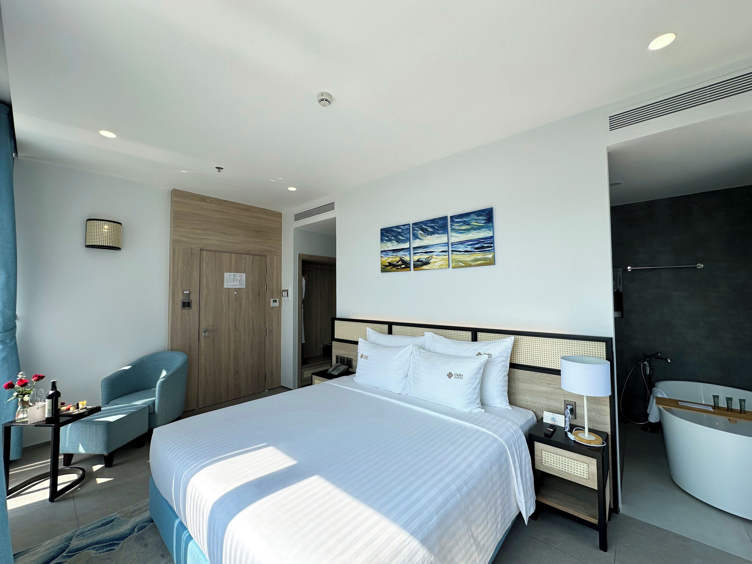 A hotel room with a bed, a chair, a desk, a nightstand, a painting, a bathroom, and a door.