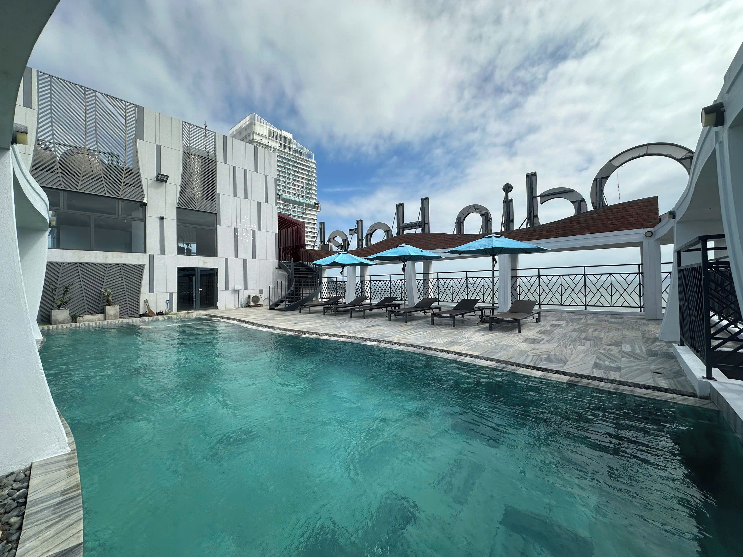 A swimming pool with lounge chairs and umbrellas, surrounded by modern buildings with a view of the sea.