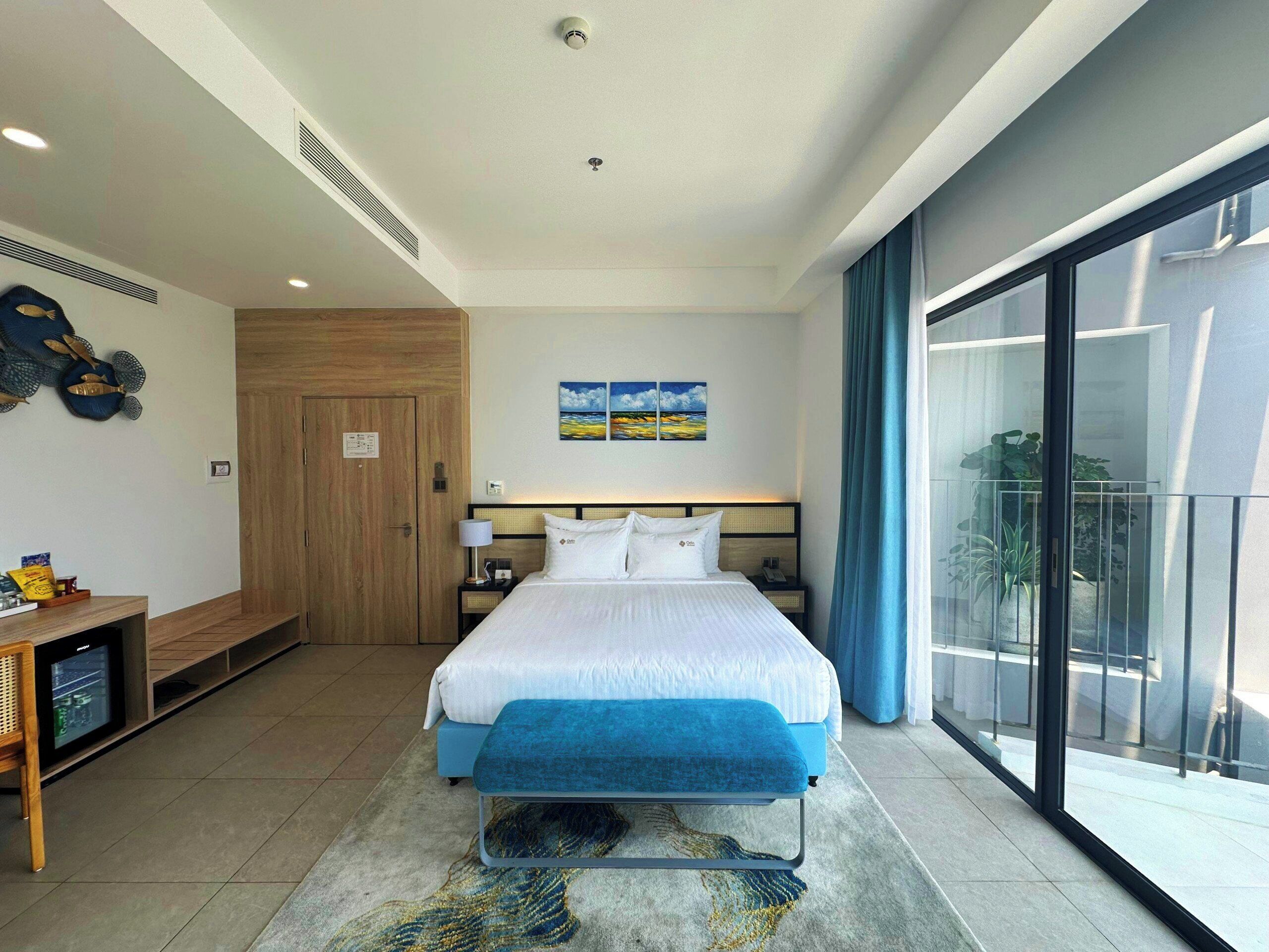 A hotel room with a bed, bedside table, lamp, artwork, and a view through a large window.