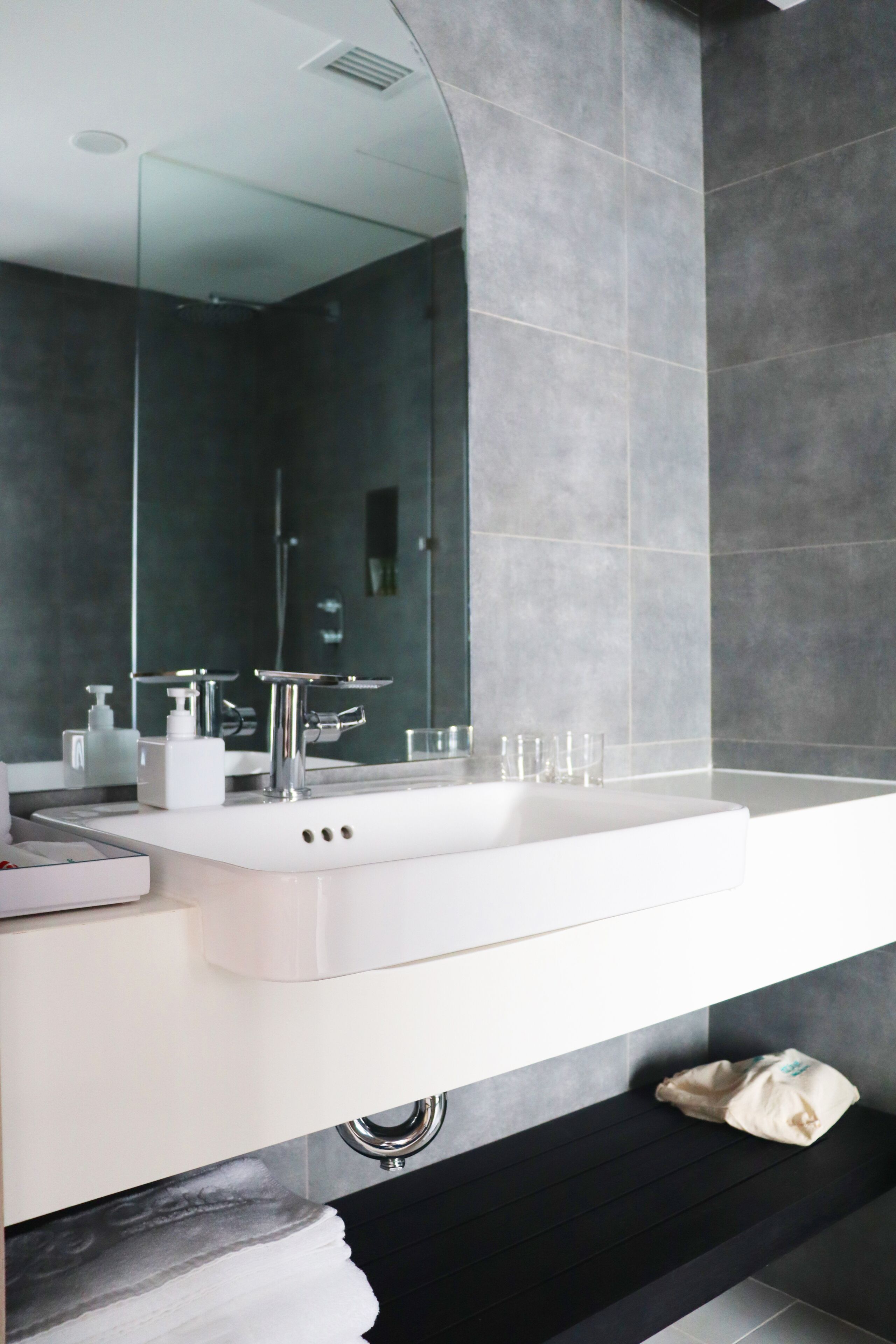 Un baño moderno con un espejo grande, lavamanos blanco y paredes cubiertas de baldosas oscuras.