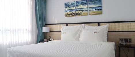 A hotel room with a large bed, white bedding, a headboard with a woven design, a wall-mounted painting of a beach scene, and a bedside table with a lamp and phone.