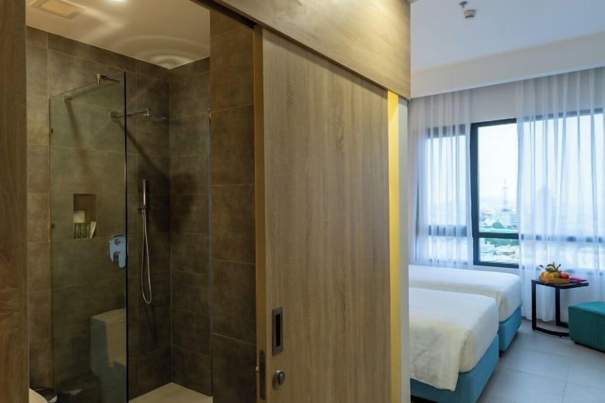 Une chambre d’hôtel dotée d’une douche en verre, d’une armoire en bois, d’un lit avec une tête de lit bleue et d’une fenêtre ornée de rideaux légers.