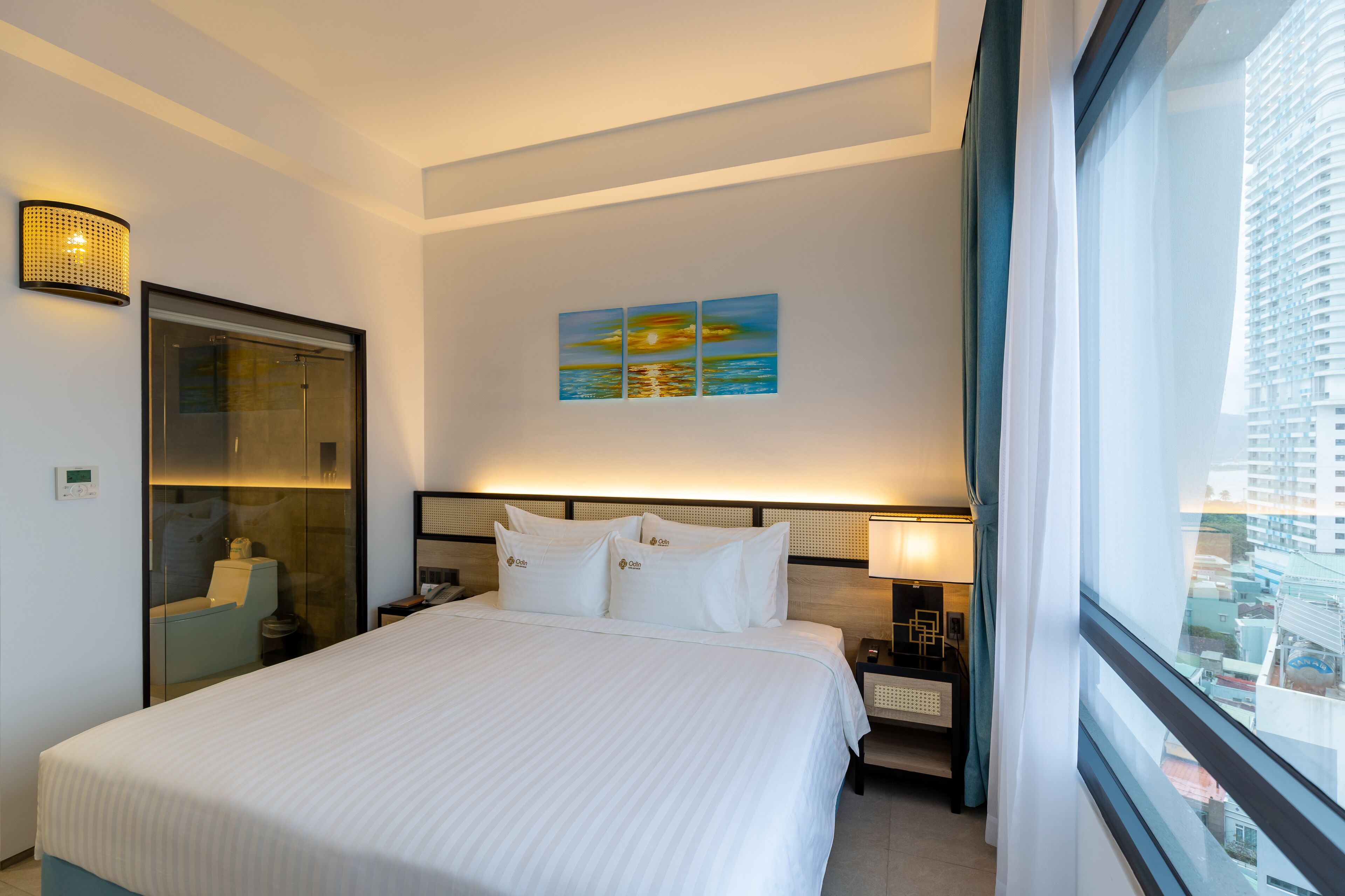 A hotel room with a large bed, a glass shower enclosure, a painting of a sunset, and a city view through the window.