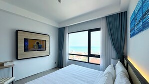 Habitación de hotel con cama, escritorio, televisión montada, ventana con vista al mar y un cuadro en la pared.