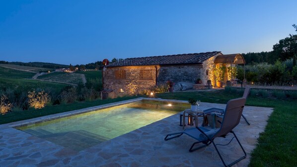 A stone house with a swimming pool, outdoor seating, and a well-lit garden at dusk.