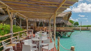 Un espace de restauration extérieur doté d’un toit de chaume, avec des chaises et des tables blanches, offrant une vue sur une eau turquoise et des bateaux.
