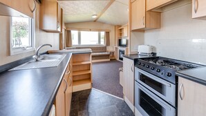 A compact kitchen with a sink, stove, and oven, wooden cabinets, and a window providing natural light. - 12ft-wide caravan with one double and two twin. (Bognor Regis)