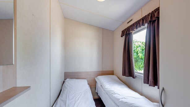 A small, single-room hotel room with two single beds, a window with curtains, and a light fixture. - 12ft-wide caravan with one double and two twin. (Bognor Regis)