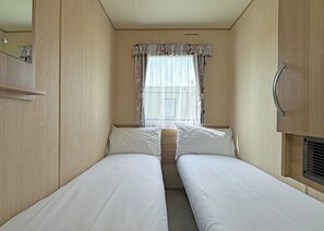 A narrow RV bedroom with two beds, a window with curtains, a mirror, and a small cabinet. - Caravan with one double and one twin. (Chapel St Leonards, Skegness)