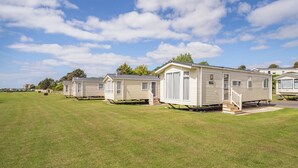 A row of mobile homes with a grassy field in the foreground and a clear sky. - 12ft-wide luxury caravan with one double and two twin. (Charmouth)