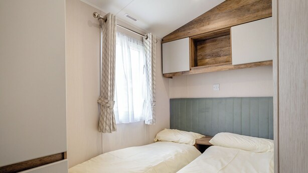 A compact bedroom with two beds, a window with curtains, a headboard, and built-in storage. - 12ft-wide luxury caravan with one double and two twin. (Charmouth)