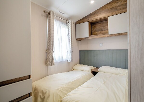 A compact bedroom with two beds, a window with curtains, a headboard, and built-in storage.
