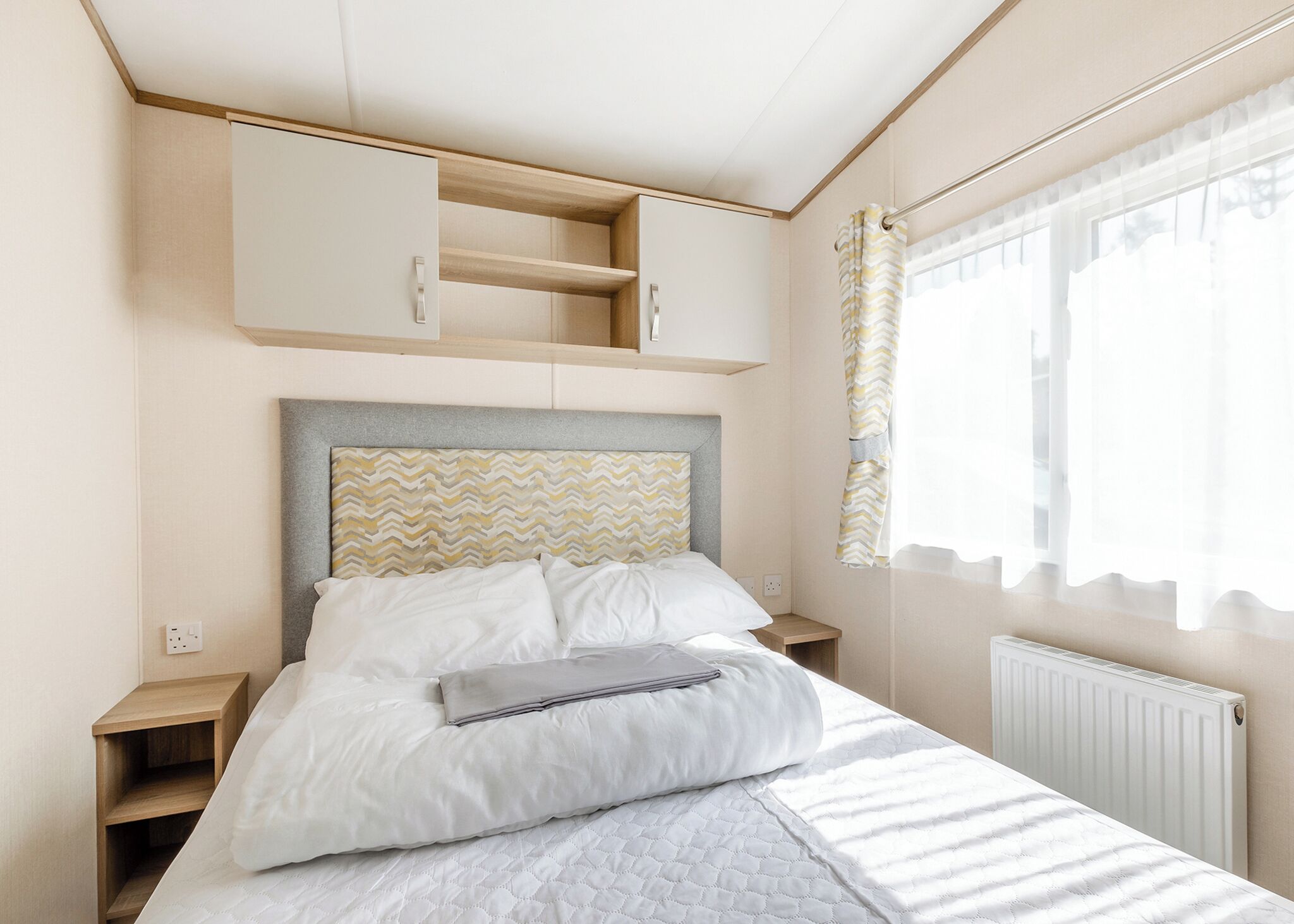 A bedroom with a bed, a headboard with a chevron pattern, a bedside table, a wardrobe, and a window with curtains.