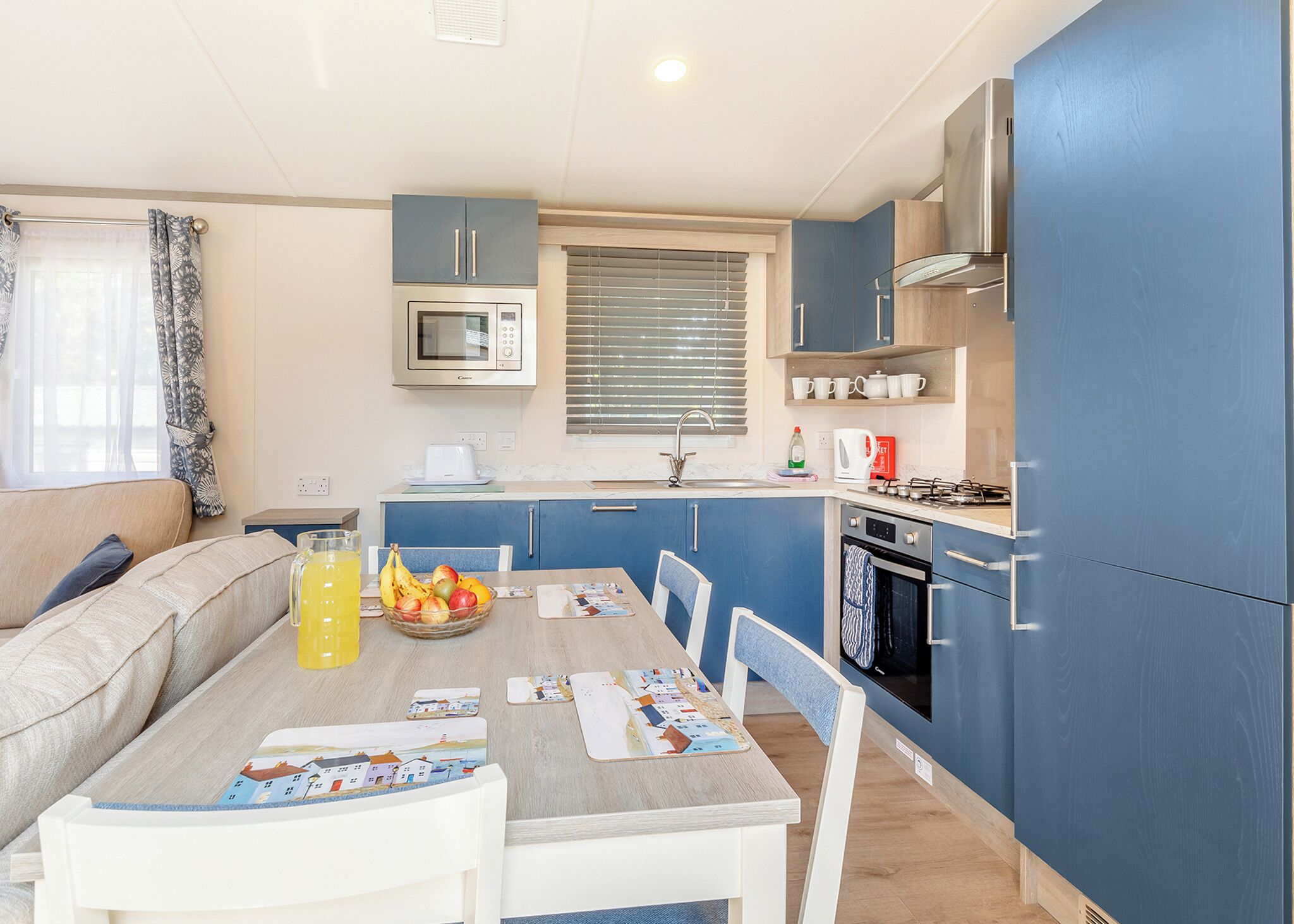 A compact kitchen with a microwave, stove, and sink. There's a dining table with chairs and a bowl of fruit.