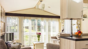 A modern kitchen with a glass-top island, a dining area with a sofa and a small table, and a view through large windows. - Oak 3 Hot Tub + Private Garden (Axminster)