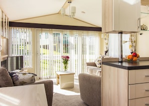 A modern kitchen with a glass-top island, a dining area with a sofa and a small table, and a view through large windows. - Oak 3 Hot Tub + Private Garden (Axminster)