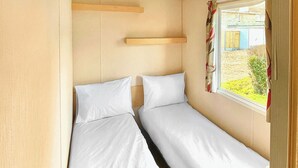 A compact room with two beds, a window with curtains, and a wooden shelf. - 3 bedroom accommodation in Newbridge, Nr Yarmouth (Newbridge, Nr Yarmouth)