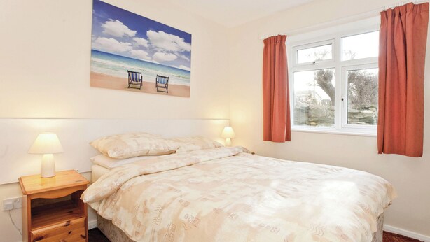 A bedroom with a bed, two bedside lamps, a nightstand with drawers, a window with curtains, and a beach-themed painting on the wall. - Bungalow with one double and one twin. (Trearddur Bay, Holyhead)