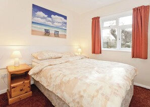 A bedroom with a bed, two bedside lamps, a nightstand with drawers, a window with curtains, and a beach-themed painting on the wall. - Bungalow with one double and one twin. (Trearddur Bay, Holyhead)