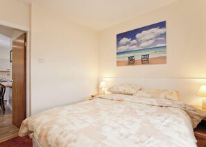 A bedroom with a bed, two bedside lamps, a painting of a beach scene, and a wooden door leading to a bathroom.