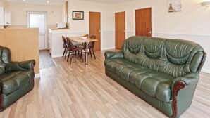A living room with a green leather sofa, wooden dining table with chairs, a kitchen area, and a door with a wooden panel. - Bungalow with one double and one twin. (Trearddur Bay, Holyhead)