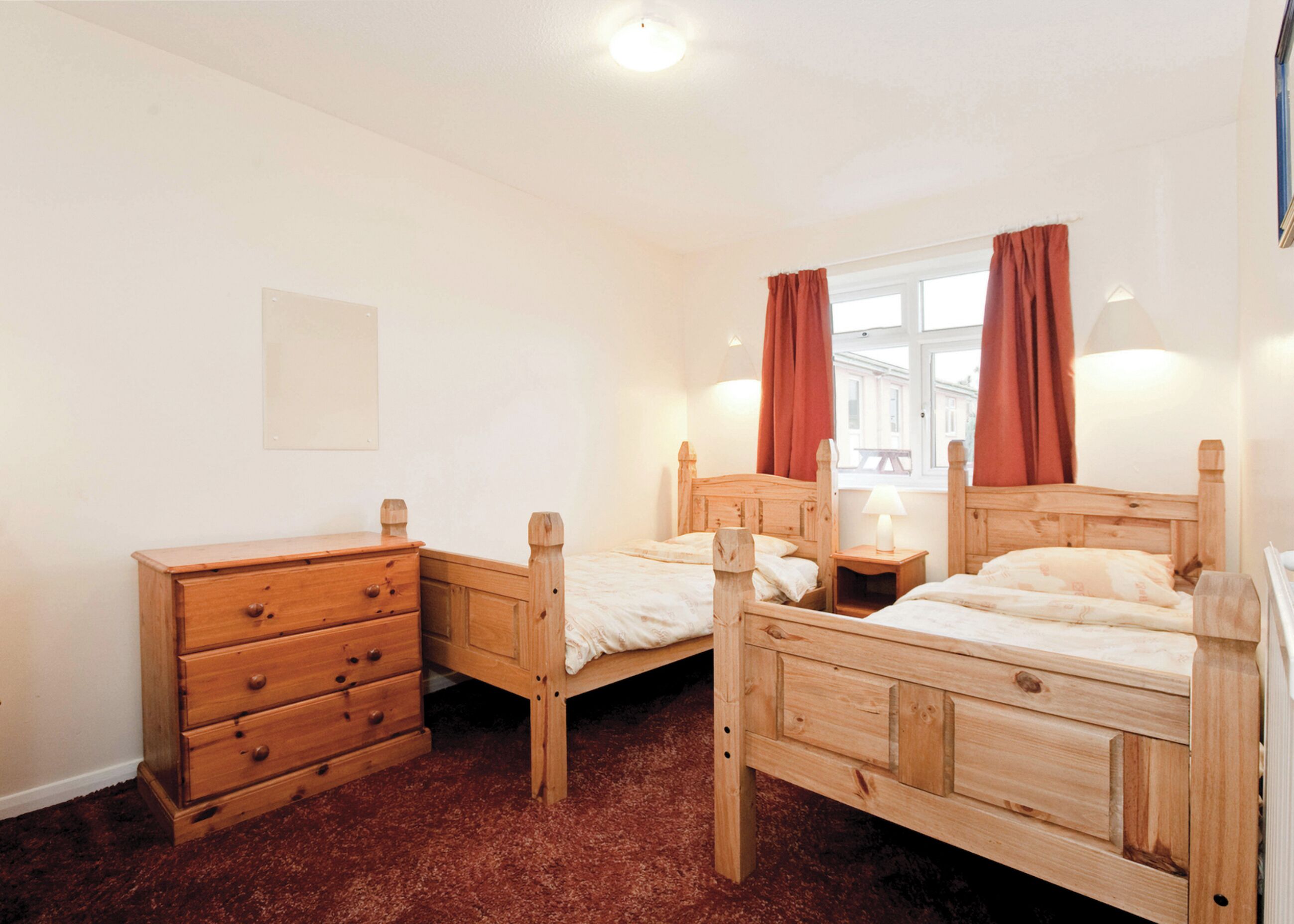 A room with two wooden beds, a wooden dresser, a window with curtains, and a wall-mounted light fixture.