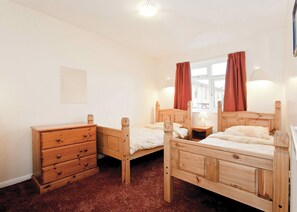 A room with two wooden beds, a wooden dresser, a window with curtains, and a wall-mounted light fixture.