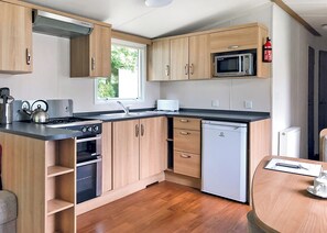 A modern kitchen with wooden cabinets, a microwave, a dishwasher, and a window providing natural light. - Luxury modern and spacious 12ft-wide caravan with one double and two twin. (Tavistock)