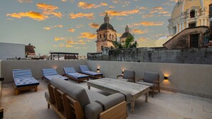 A rooftop terrace with lounge chairs, a wooden table, and a view of historic buildings against a sunset sky. - 5 Bedroom Luxury Villa in Old City (Cartagena)