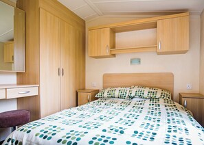 A bedroom with a bed, a nightstand, a dresser, a mirror, and a small stool. - Well presented 12ft-wide caravan with one double and one twin. (Clunderwen, Nr Narberth)