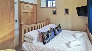 Un letto rifatto con lenzuola bianche e cuscini decorati, una testiera in legno, una porta di legno, una piccola finestra e una TV a schermo piatto montata a parete.